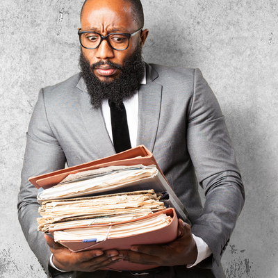 A man wearing a gray suit and glasses holding a large stack of business documents looking at them with concern.