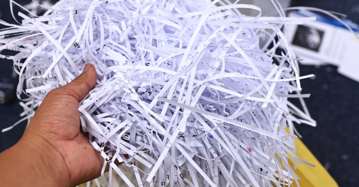 A person’s hand holding out the remains of shredded documents over a yellow recycling container in an office.
