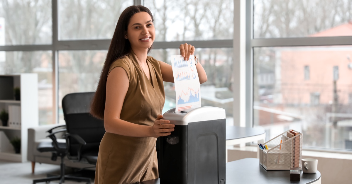 A woman wearing a tan dress standing in an office while feeding a document through a shredding machine.