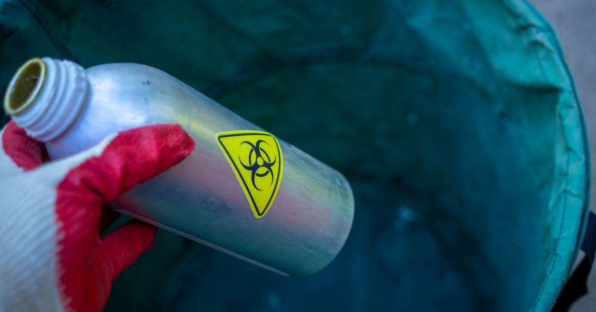A person wearing a glove is disposing of a container with a biohazard sticker on it in a green bin.