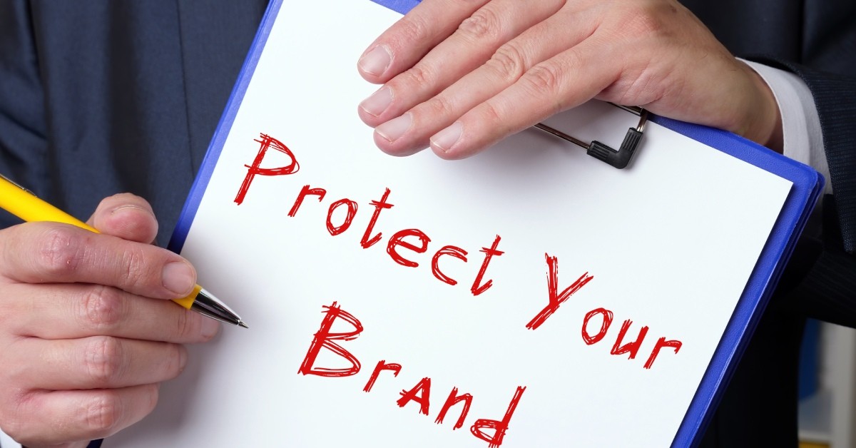 A person wearing a suit holds a pen and a clipboard with the words "Protect Your Brand" written in red.