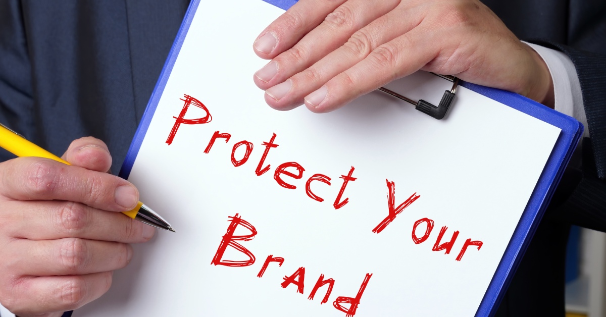 A person wearing a suit holds a pen and a clipboard with the words "Protect Your Brand" written in red.