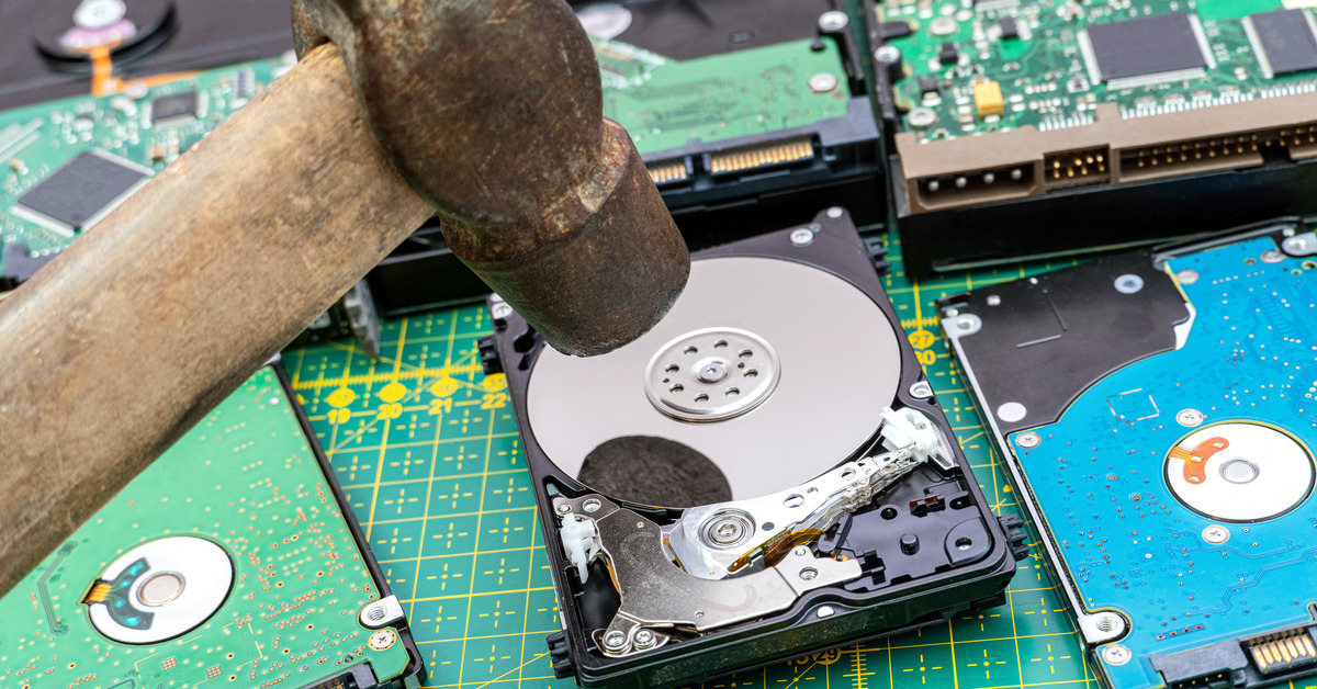 Several hard drives sit on a table. Someone is holding a hammer over one of them and getting ready to smash it.
