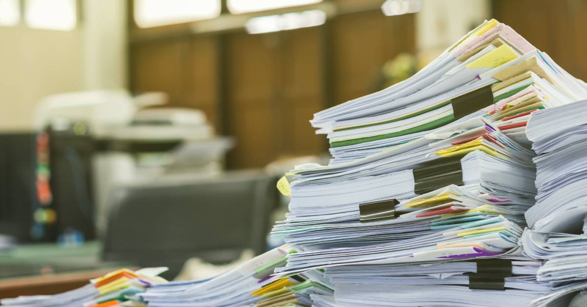 Multiple piles of business documents sitting on a desk in an office. There is a chair and a printer in the background.