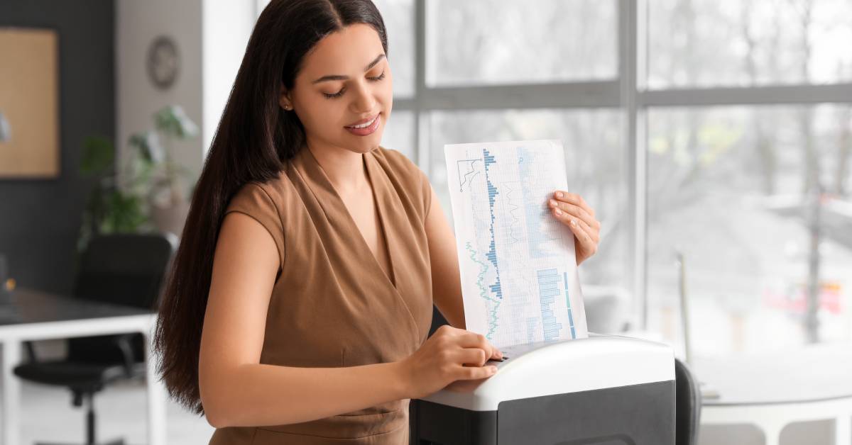A woman in a beige outfit standing in an office near some windows. She is feeding a document through a shredder.