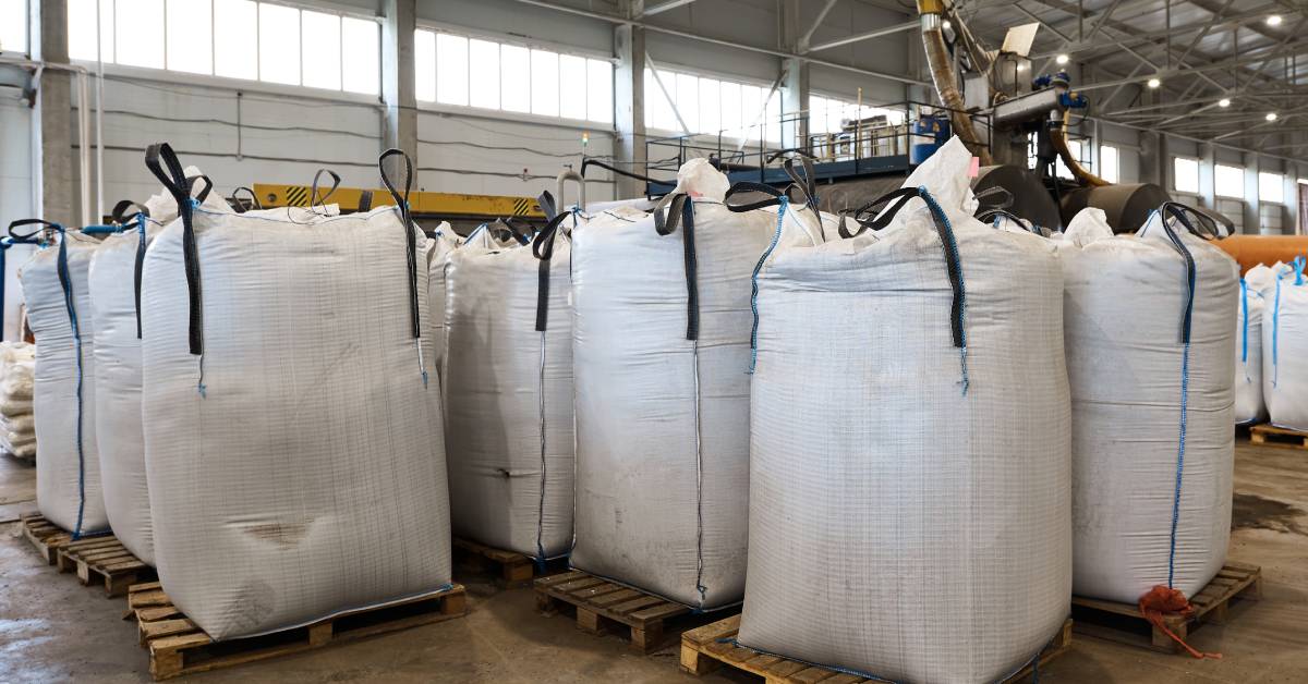 Large white bags filled with products sitting on wooden pallets in a warehouse with equipment operating behind them.