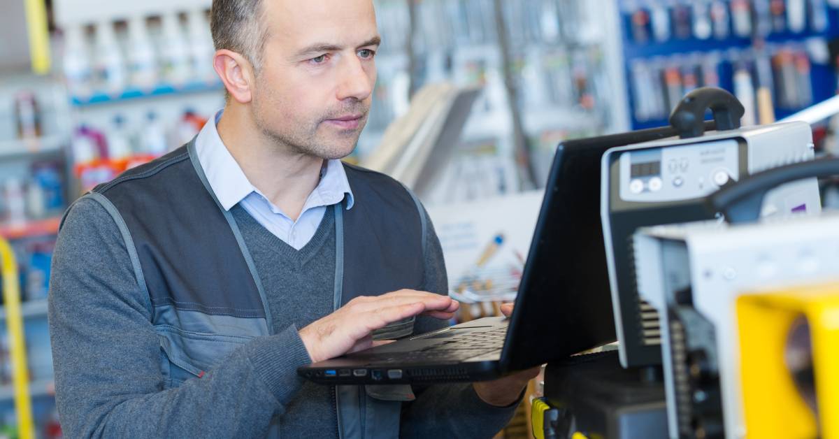 A man wearing a long-sleeve shirt standing in a product disposal warehouse while operating a computer.