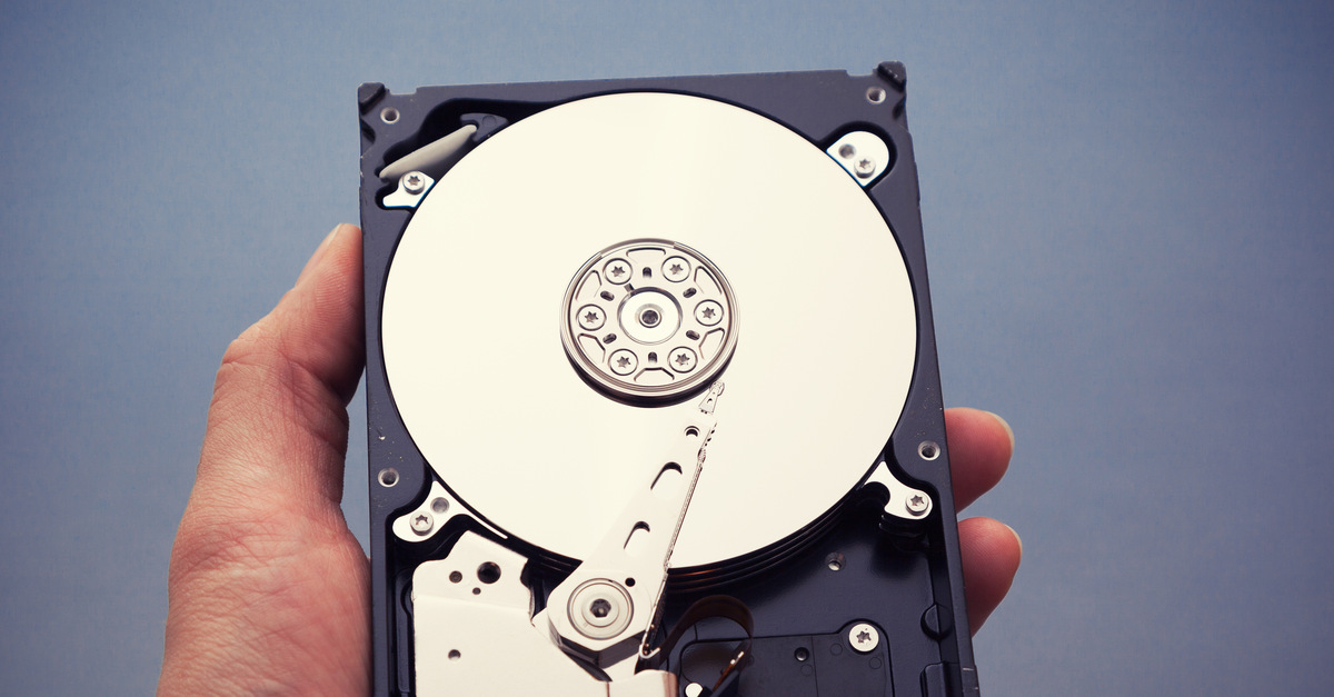 A person’s hand holding a physical hard drive with the moving parts facing upwards over a gray background.