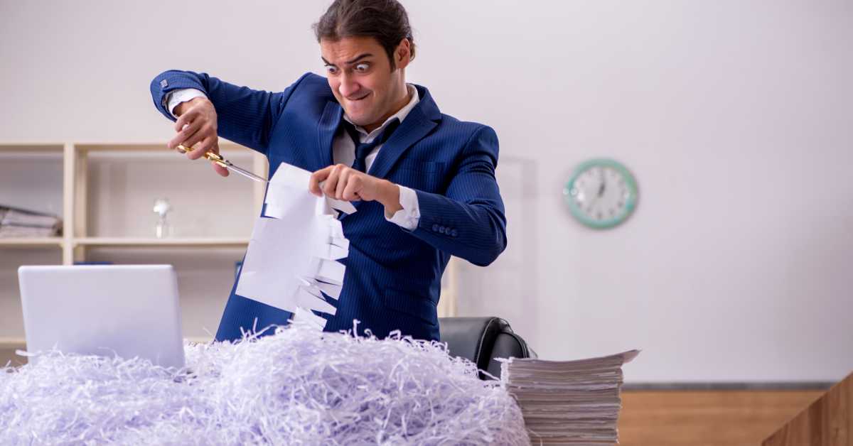 A man with a disturbed look wearing a blue suit standing in front of his desk in an office using scissors to cut a document.