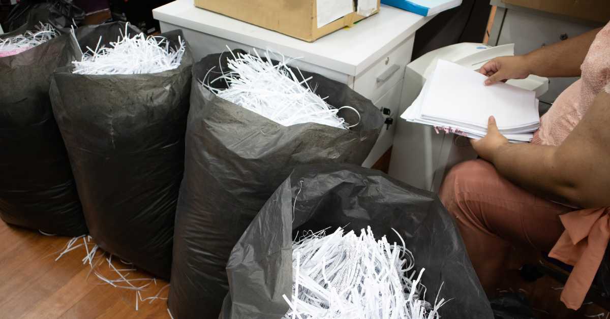 A woman sitting at a desk in an office using a machine to shred documents with the remains in trash bags.