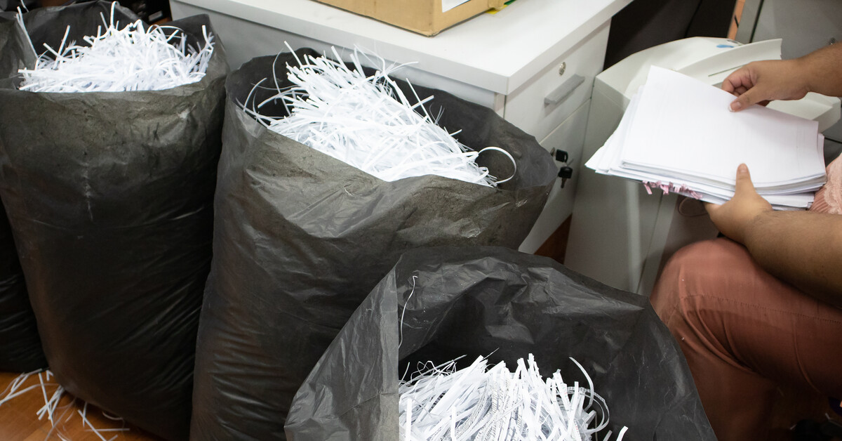 A person sitting at a desk in an office in the process of shredding old documents into large black trash bags.