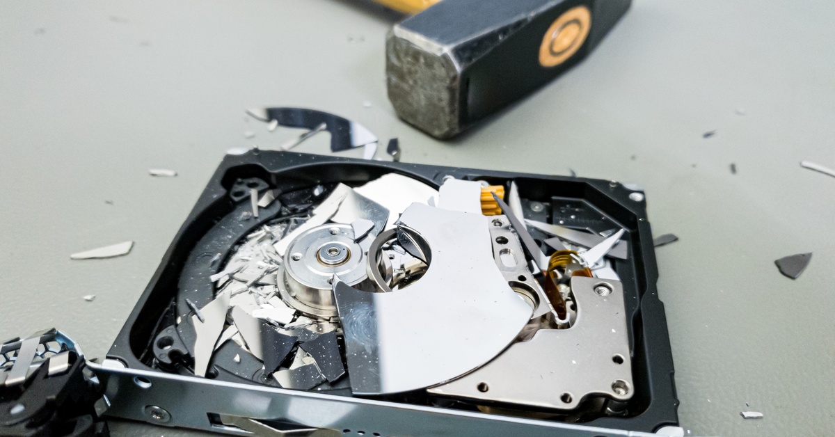 A hammer with a wooden handle laying next to a hard drive that’s been smashed into small pieces on a cement floor.
