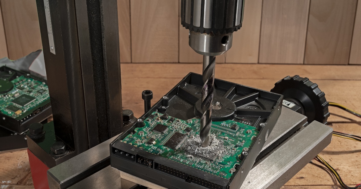 A computer hard drive on a bench with a drill press being driven into it. There are more hard drives stacked next to the press.
