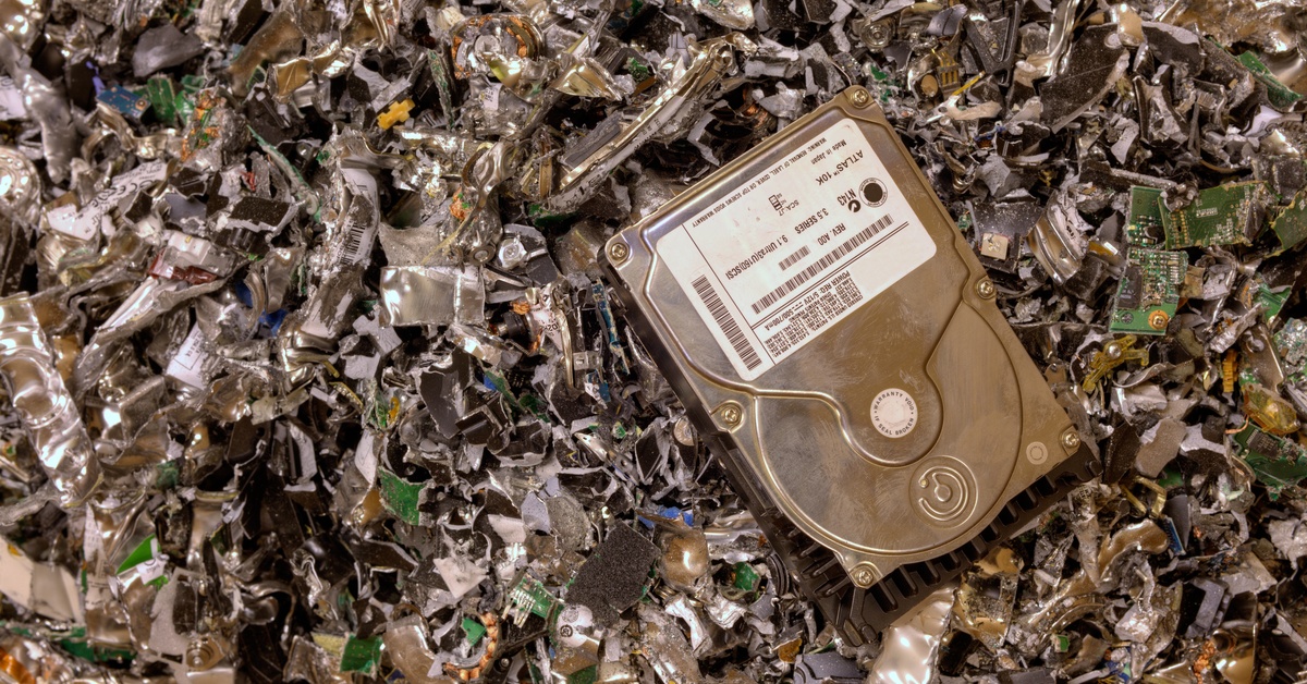 An intact hard drive with a white label on the casing sitting on top of a pile of shredded electronic drives.