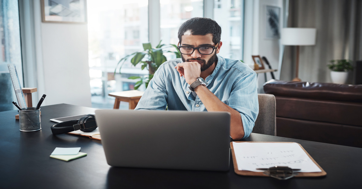 A man sitting at a desk in his home office thinking about something while using the computer in front of him.