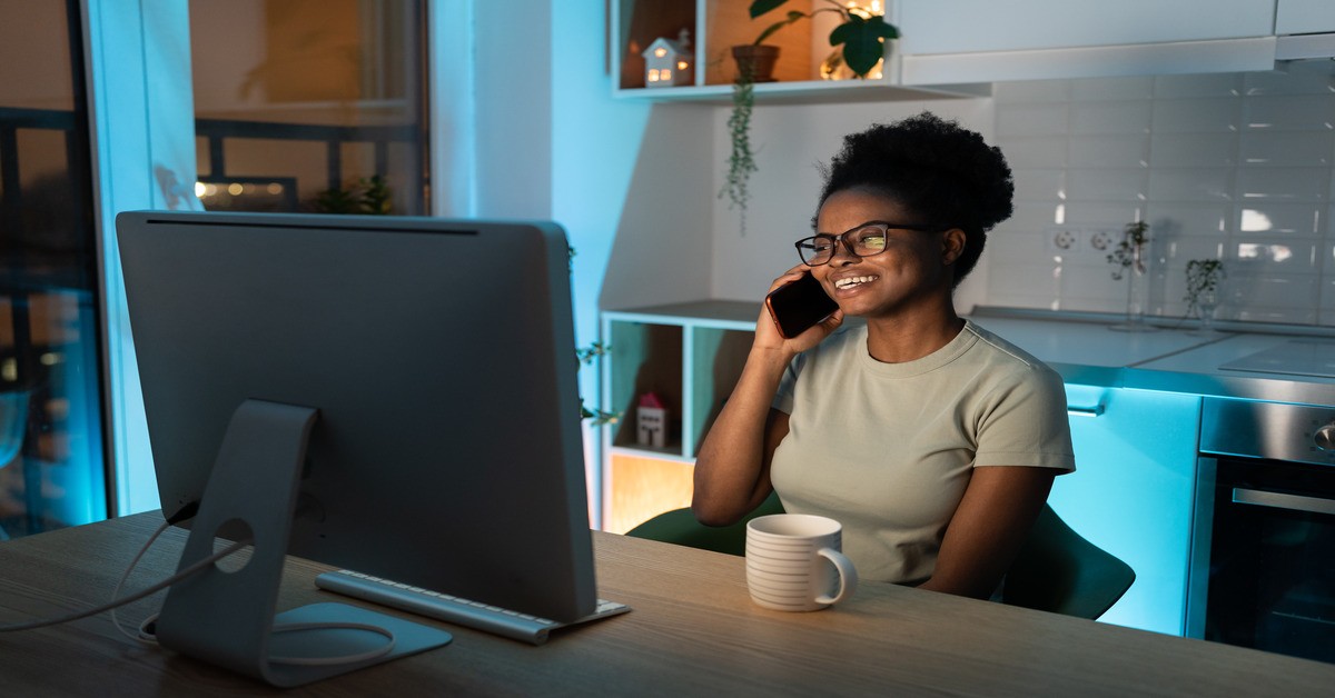 A woman sitting at a desk in her home office with a computer in front of her while talking on the phone with someone.