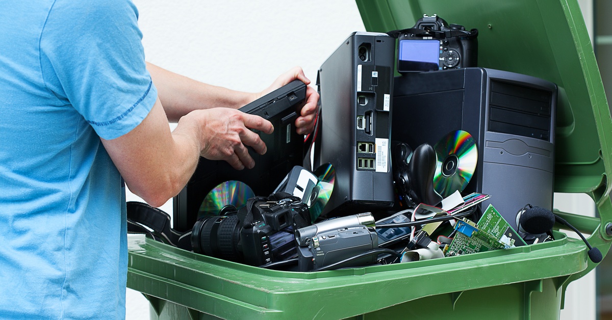A man wearing a blue shirt placing old unwanted electronics in a green recycling bin in front of a white wall.