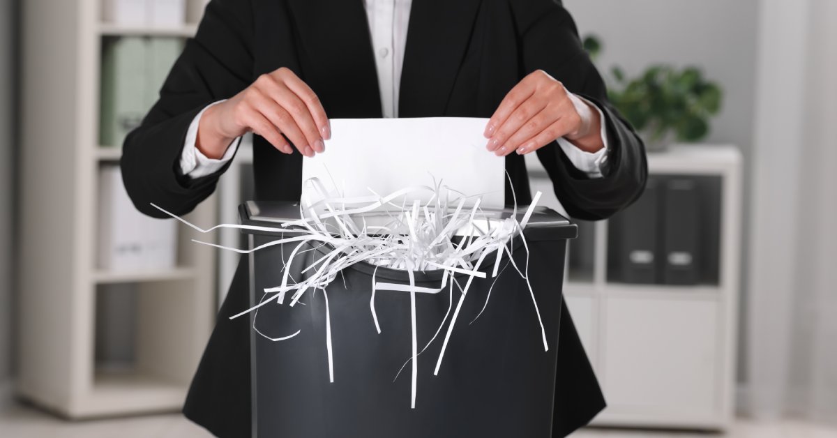 A person in a black suits stands in an office and feeds a piece of paper into a shredder that is overflowing.