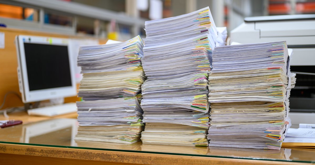 Three tall stacks of paper that are waiting to be disposed of sit on a desk in an office next to a computer.