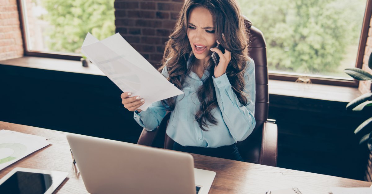 A woman in an office sits at a desk in front of a computer while holding up a document and talking on the phone.