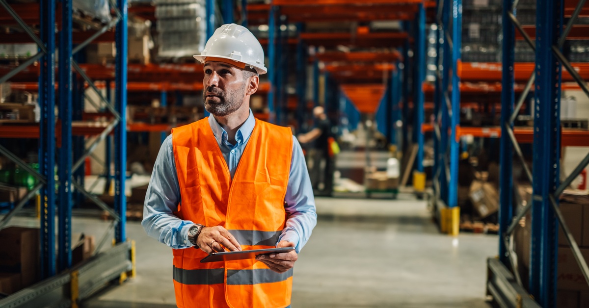 A worker in a warehouse wearing a hard hat and high-visibility vest holds a clipboard while inspecting the area.