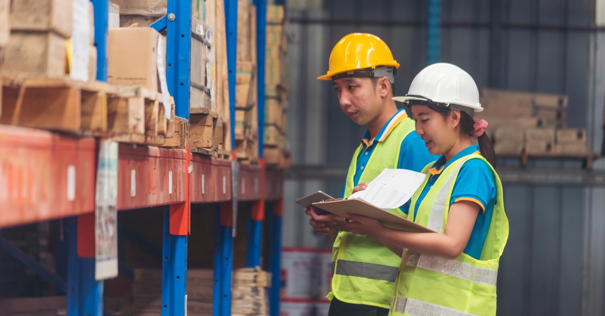 Two workers wearing hard hats and high-visibility vests looking through the stock in a warehouse while taking notes.