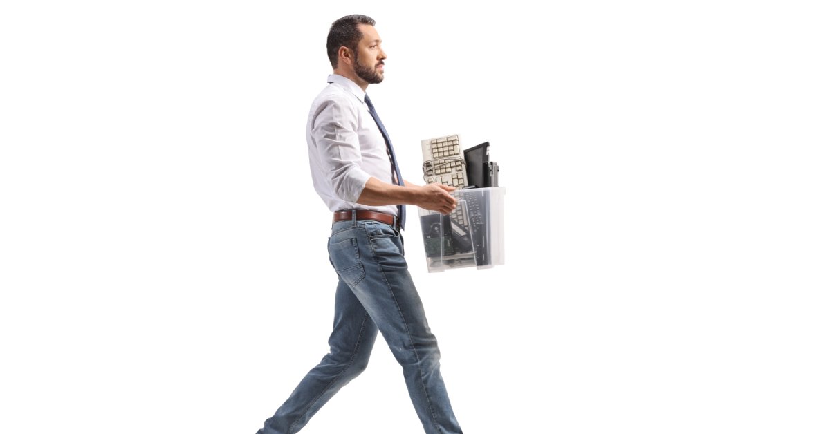 A man wearing a button-down shirt and tie walking with a box of old electronics in front of a white background.