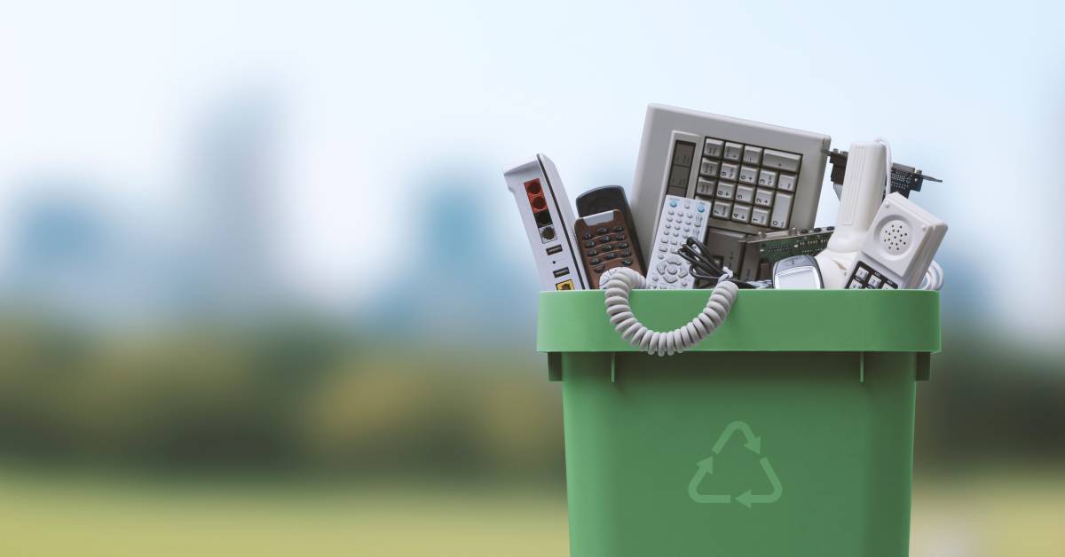 A green recycling container with various discarded electronics inside of it in front of a blurry background with trees.