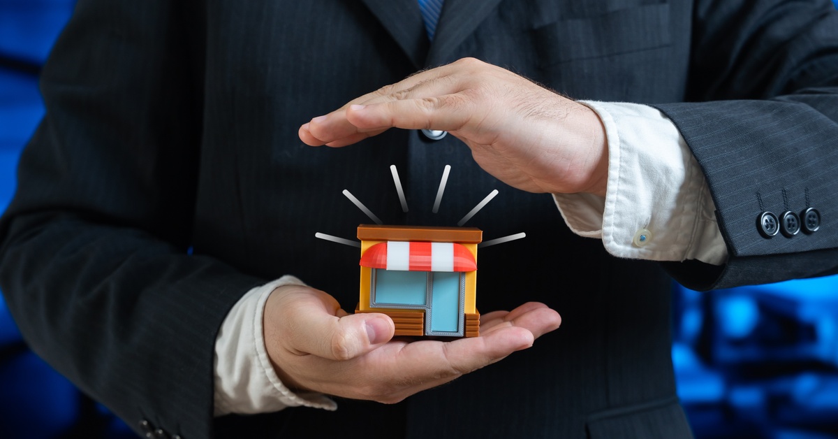 A business person cupping a small toy storefront in one hand and protectively shielding it from above with the other.
