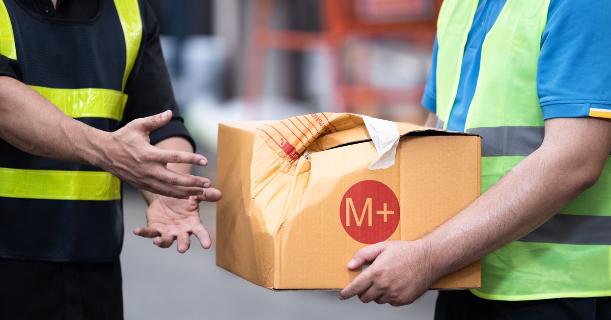 A warehouse worker handing off a branded cardboard box. The box has a large dent in the side with crumbled material.