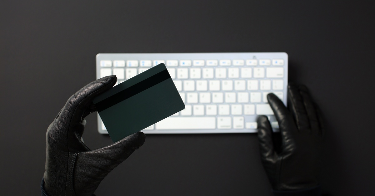 A gloved hand holding a simple credit card toward the viewer. A white keyboard is visible in the background.