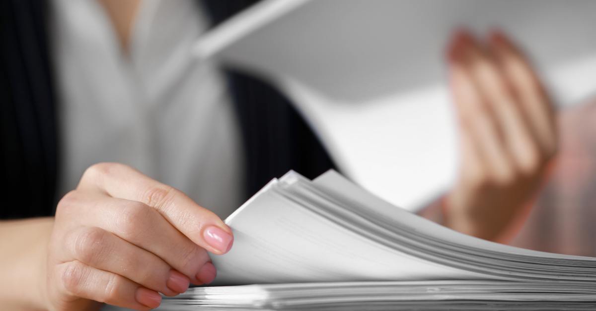 A person in professional attire sorting through a large stack of white documents. She holds one half in her hand.