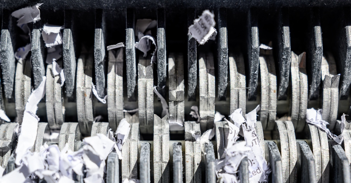 A close-up on the teeth of an industrial shredder that is tearing apart shreds of paper with black text on them.