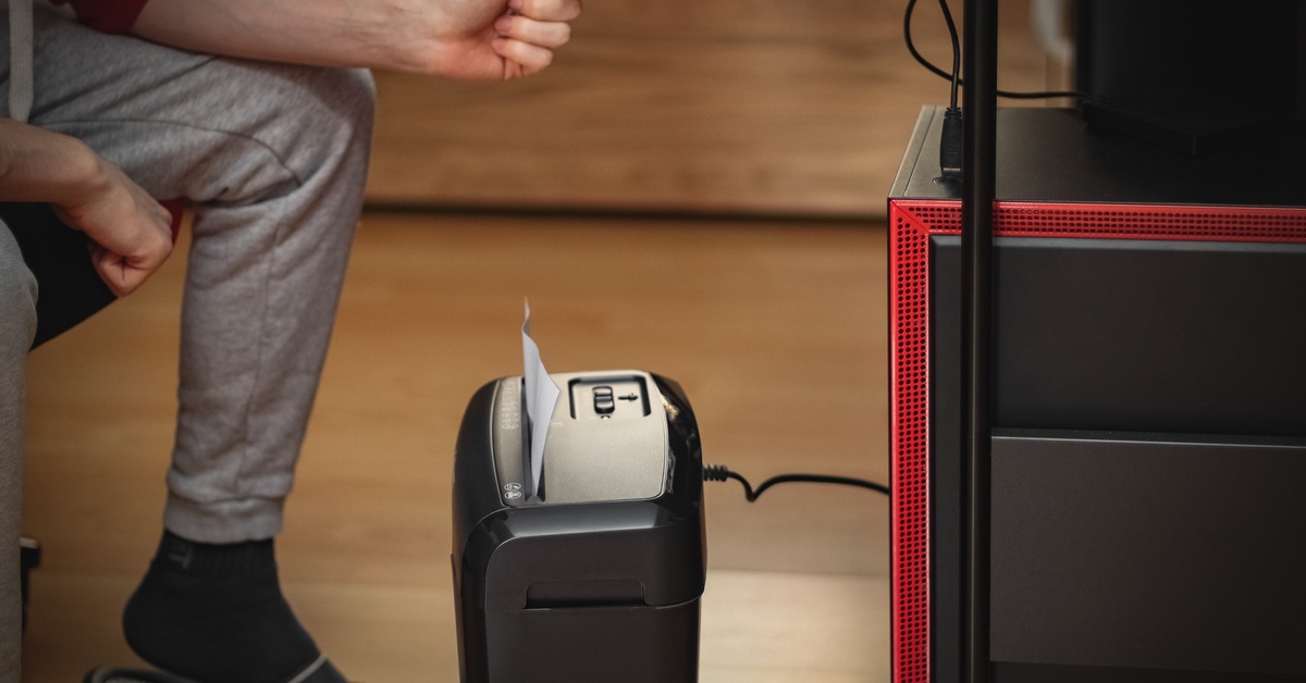 A person sitting at their home desk and watching as a personal shredder on their floor shreds a white paper document.