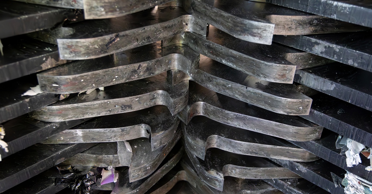 The underside of an industrial shredder. Large metallic teeth move in an interwoven design to shred paper pieces,