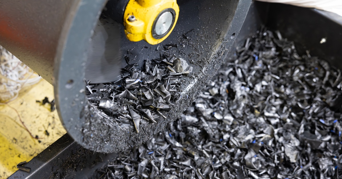 A recycling plant machine processing shreds of dark gray plastic waste into a large black collection bin.
