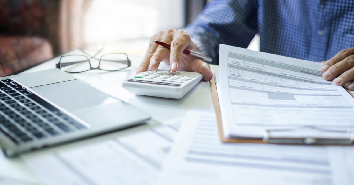 A person sitting at a table and reviewing accounting and tax documents while using a small calculator.