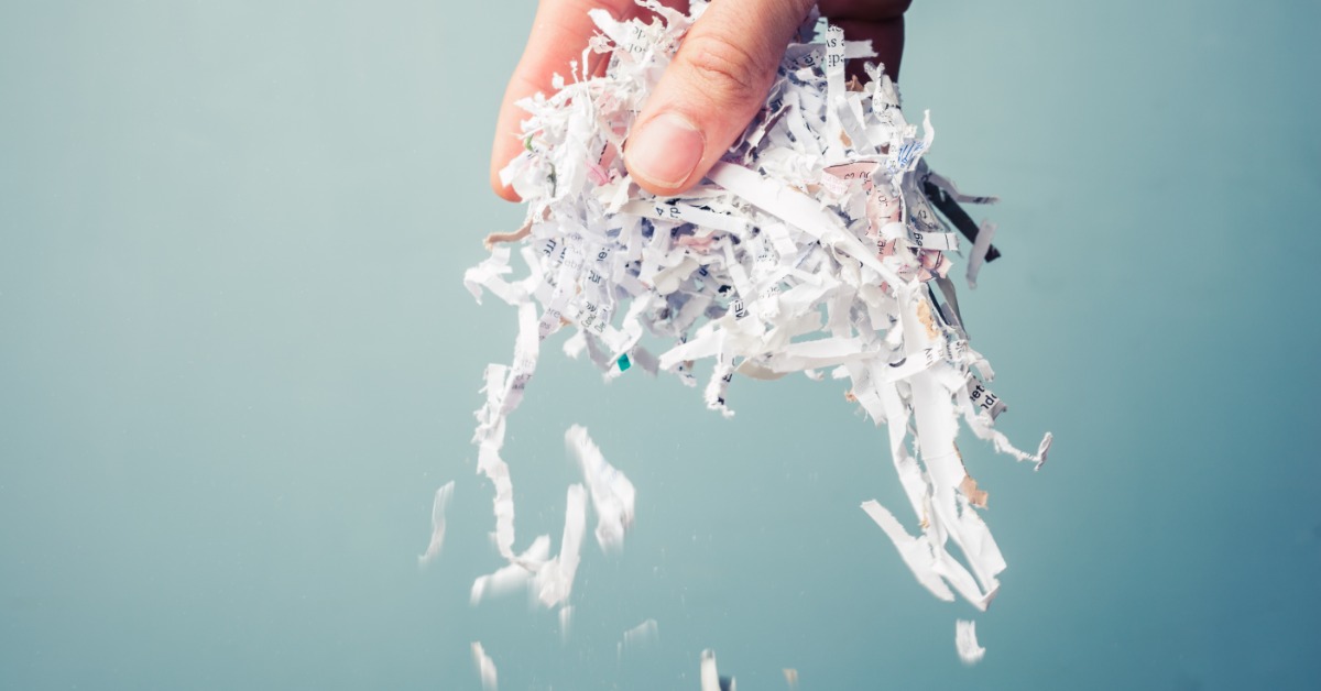 A hand lifting a wad of paper shreds with limited visible text on them. A few paper shreds shower down from the hand.