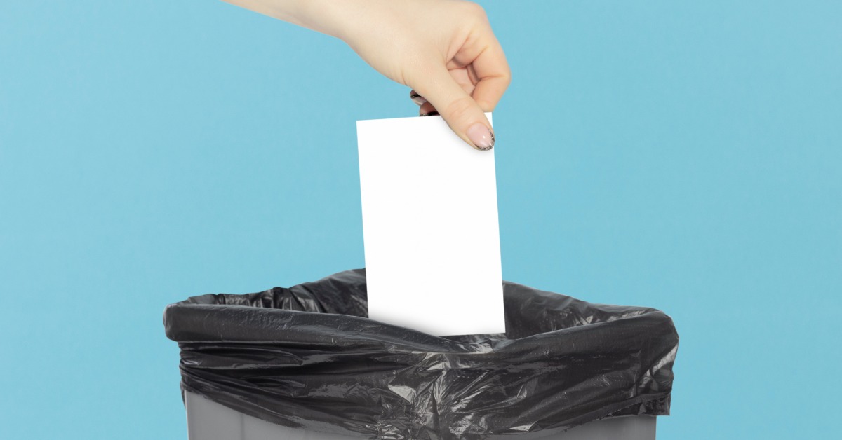 A person with manicured nails lowering a white piece of paper into a trash bin bag against a blue background.