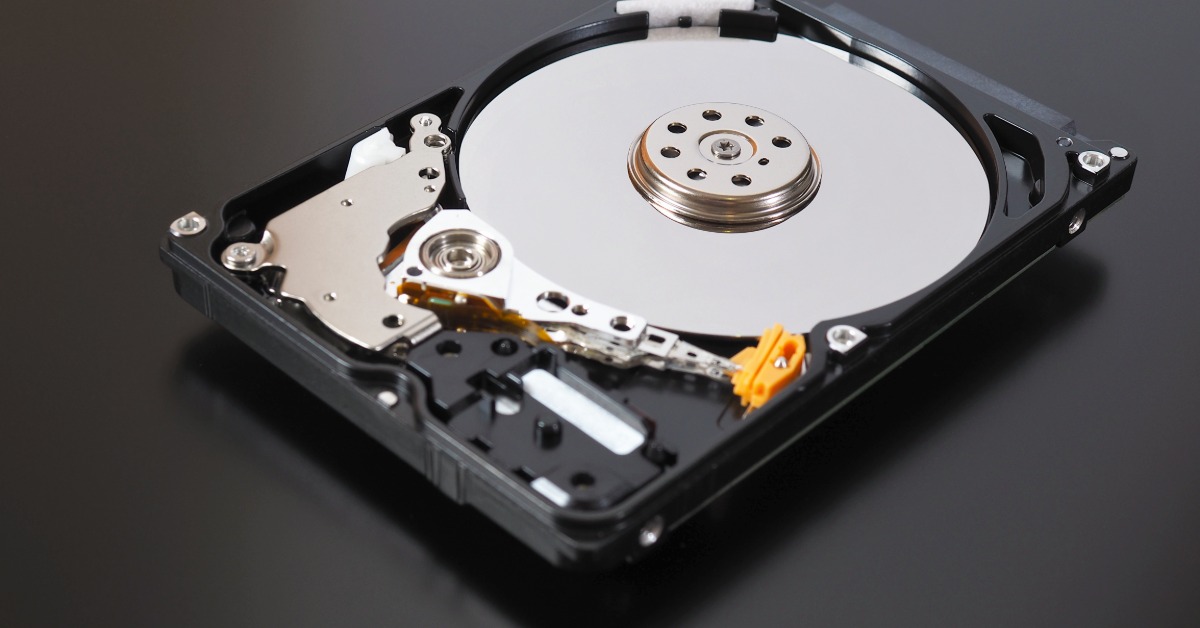 An open hard disk drive rests on a dark surface with the silver platter, actuator arm, screws, and black casing visible.