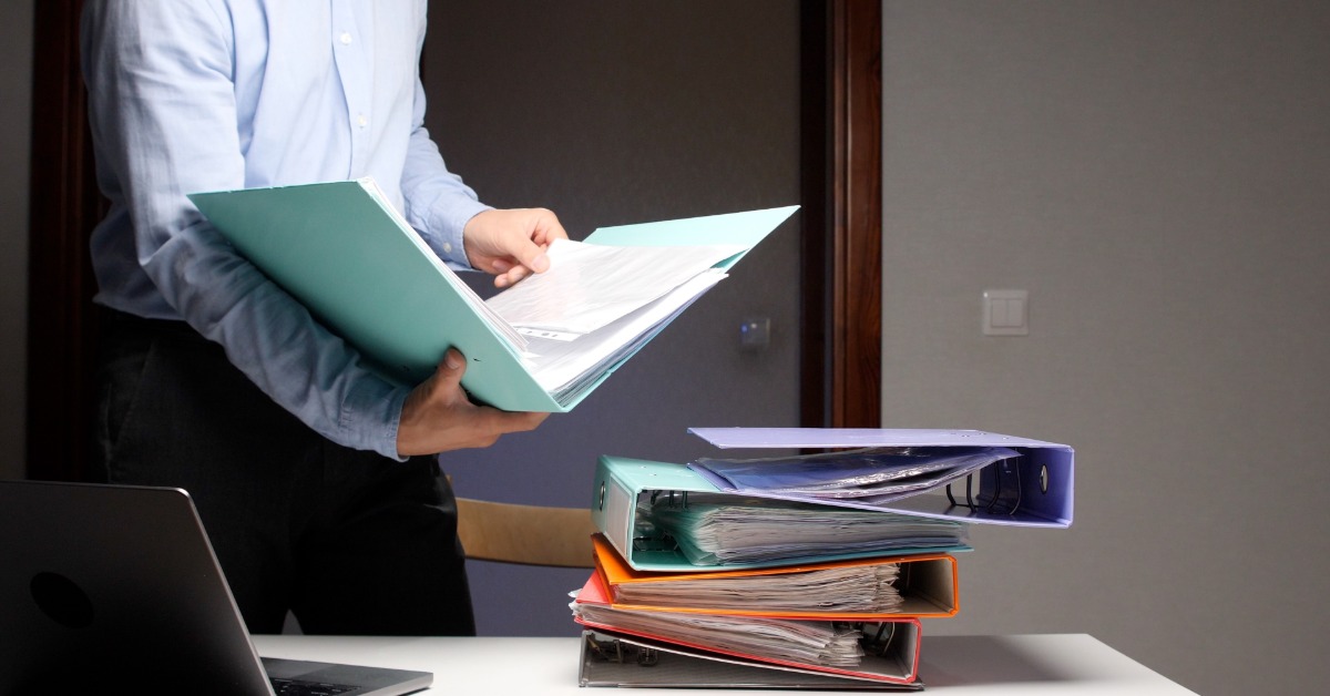 An office worker reviews documents in a large teal binder. More binders and a laptop are on the tabletop next to him.
