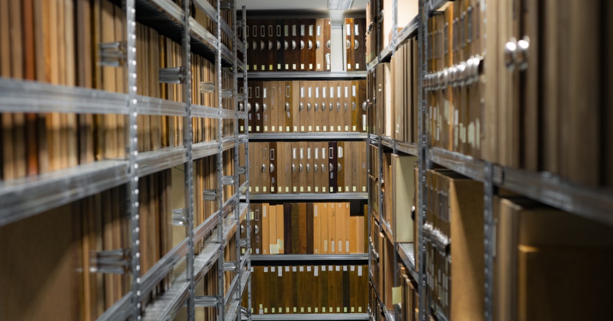 Rows of metal shelving hold cardboard archive binders on both sides of a narrow aisle and at the far end of the room.