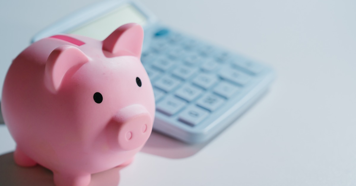A pink piggy bank sits on a white surface in front of a calculator. The calculator is slightly blurred.