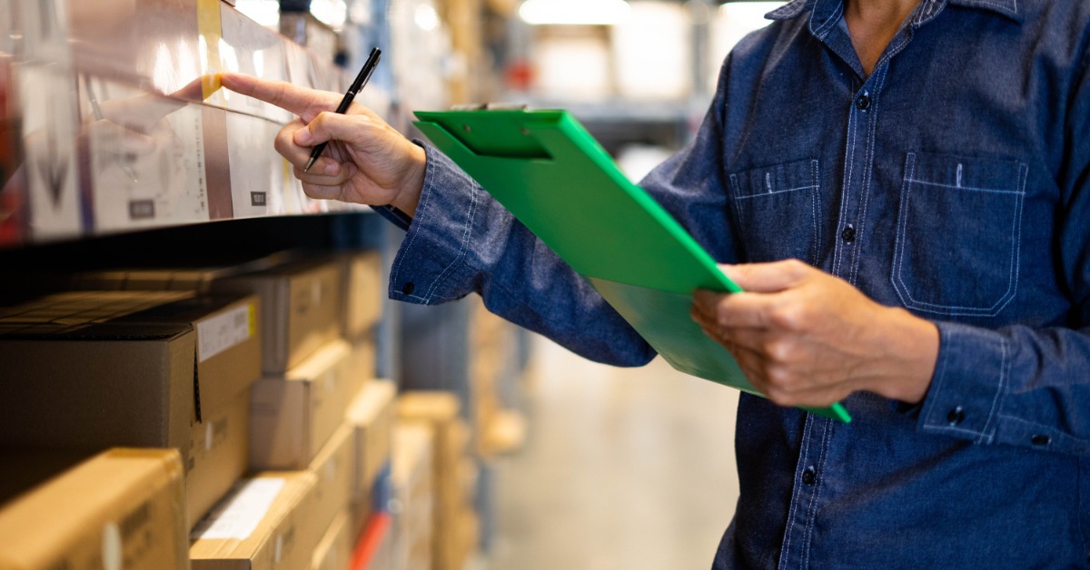 A worker in a warehouse holds a green clipboard and stands by shelves of stacked boxes. They point at a label on a box.