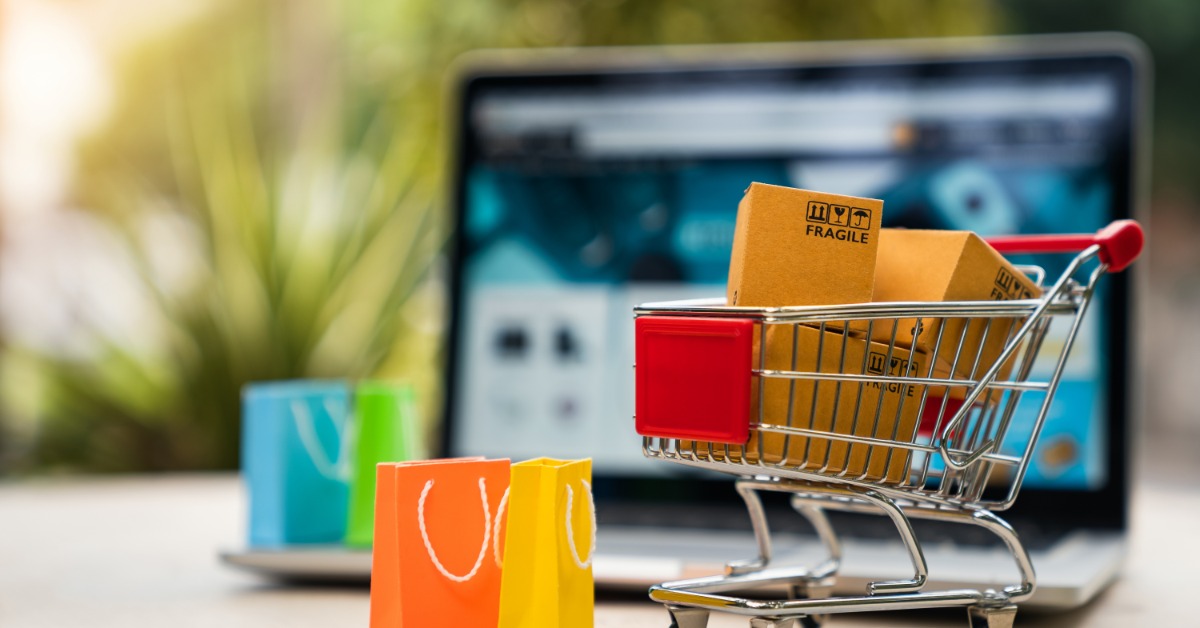 In front of a laptop, a miniature shopping cart holds cardboard boxes. Miniature shopping bags are near the cart.
