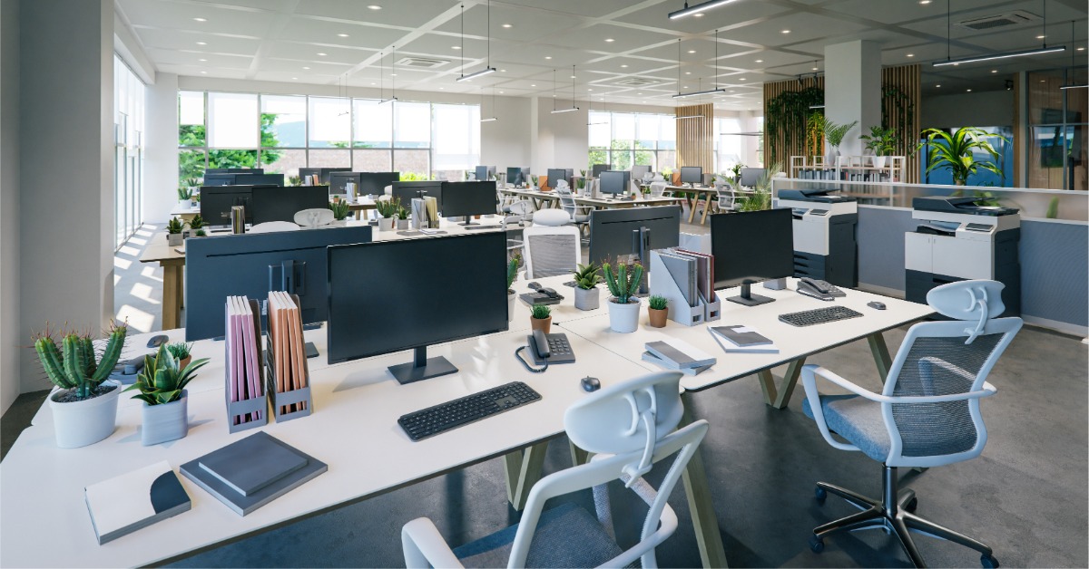 A bright open-plan office contains rows of white desks with black computer monitors, keyboards, and office supplies.