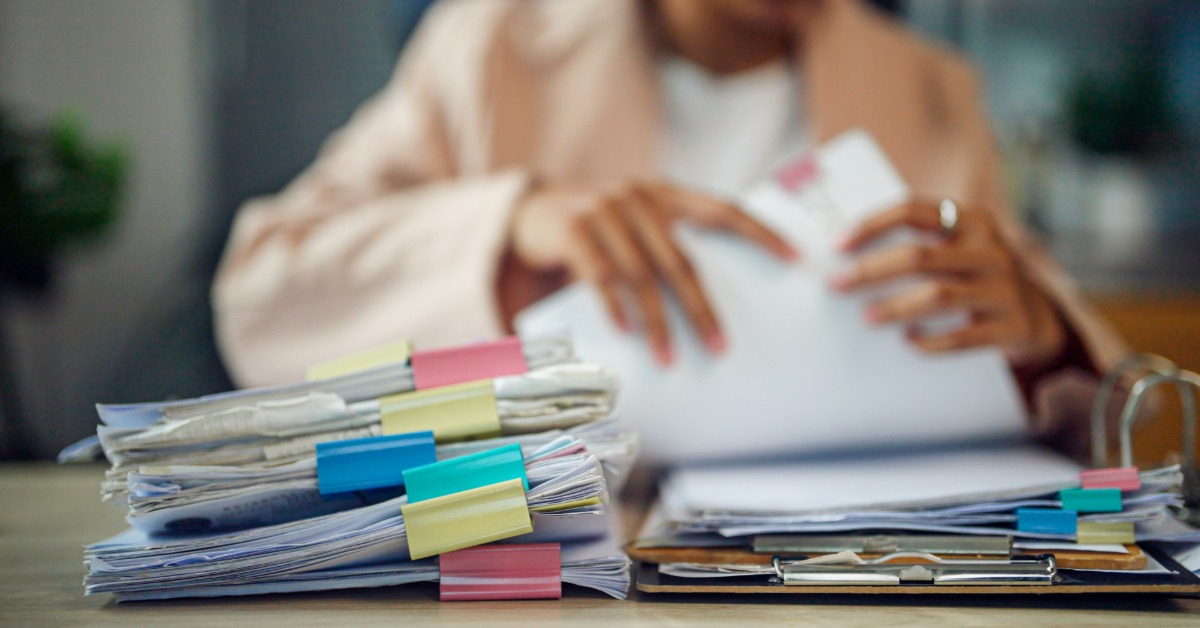 A person in a blazer sorts through piles of paperwork held together with pink, blue, yellow, and teal binder clips.